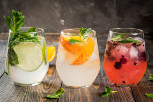 Selection of three kinds of gin tonic: with blackberries, with orange, with lime and mint leaves. In glasses on a rustic wooden background. Copy space